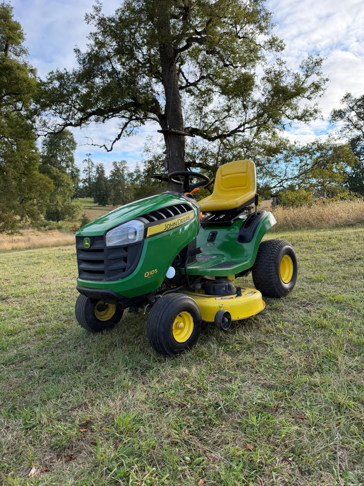 Vista frontal lateral de un tractor cortacésped verde y amarillo bajo un árbol.